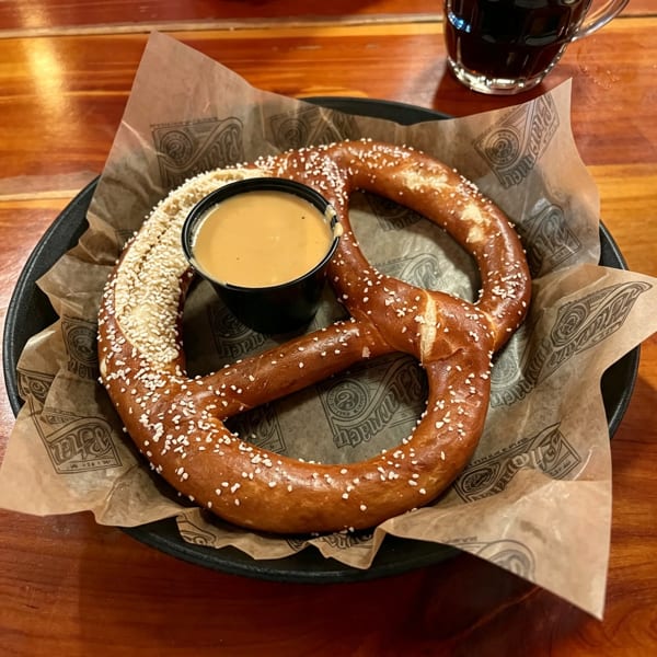 A large, salted pretzel with a side of cheese dip on branded parchment paper, with a dark drink in the background.