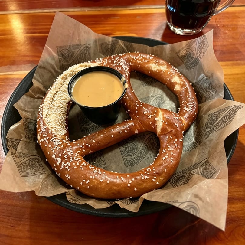A large, salted pretzel with a side of cheese dip on branded parchment paper, with a dark drink in the background.