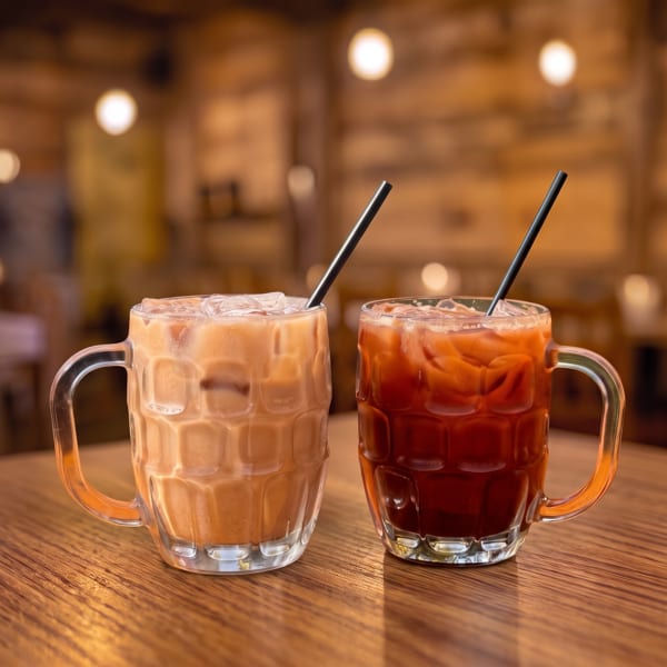 Two iced Thai teas, one creamy and one dark, in glass mugs with straws on a wooden table.