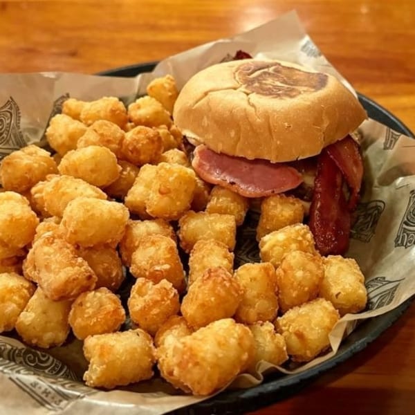 A burger with bacon and a side of crispy tater tots on a black plate with branded paper.