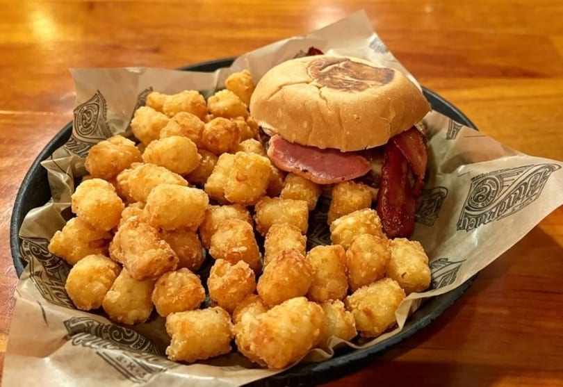 A burger with bacon and a side of crispy tater tots on a black plate with branded paper.