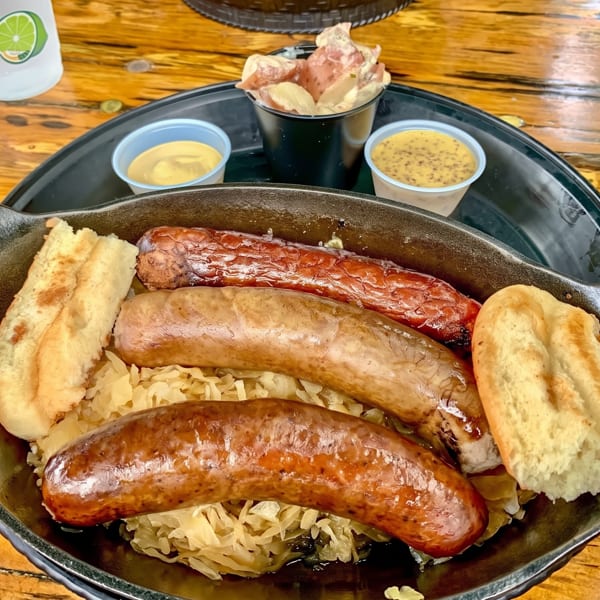 Three sausages, sauerkraut, and bread in a black skillet, with sides of mustard and potato salad.