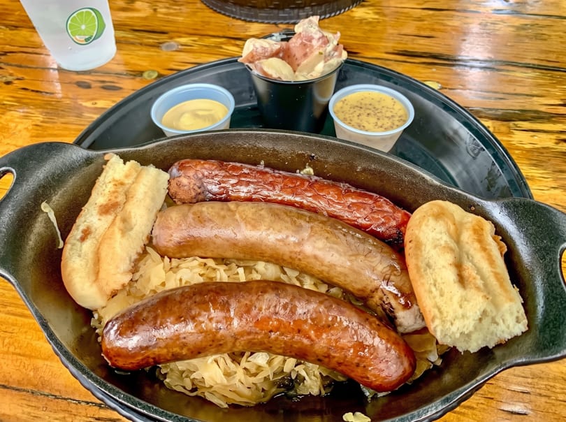 Three sausages, sauerkraut, and bread in a black skillet, with sides of mustard and potato salad.